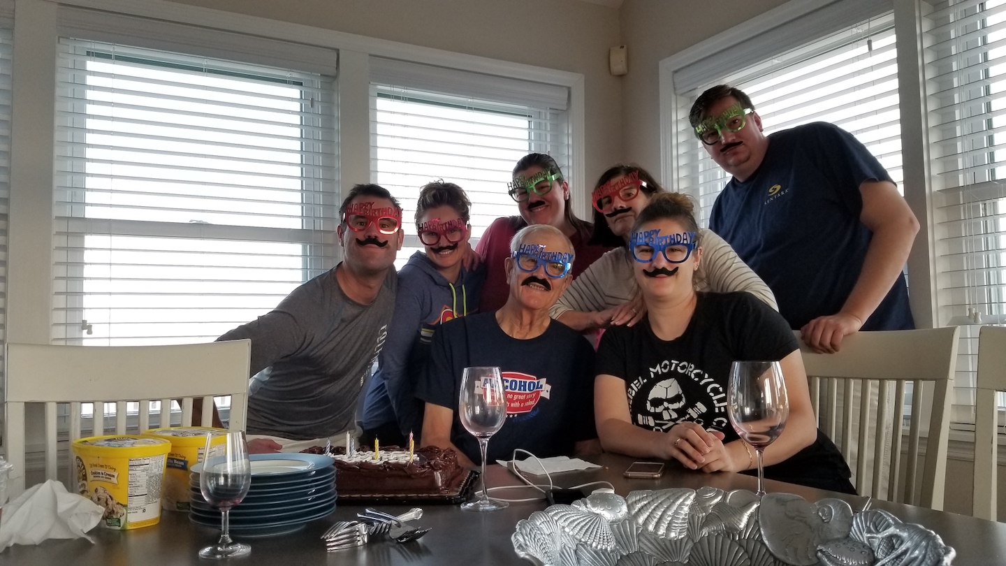 A family of white folks all wearing novelty paper glasses with the words Happy Birthday on them and mustaches hanging below smile at the camera while the remnants of a cake sit on the table in front of them