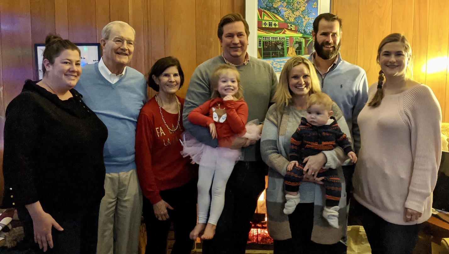 A family of white people, seven adults and two small children, stand against a wood panelled wall and pose for a family photo.