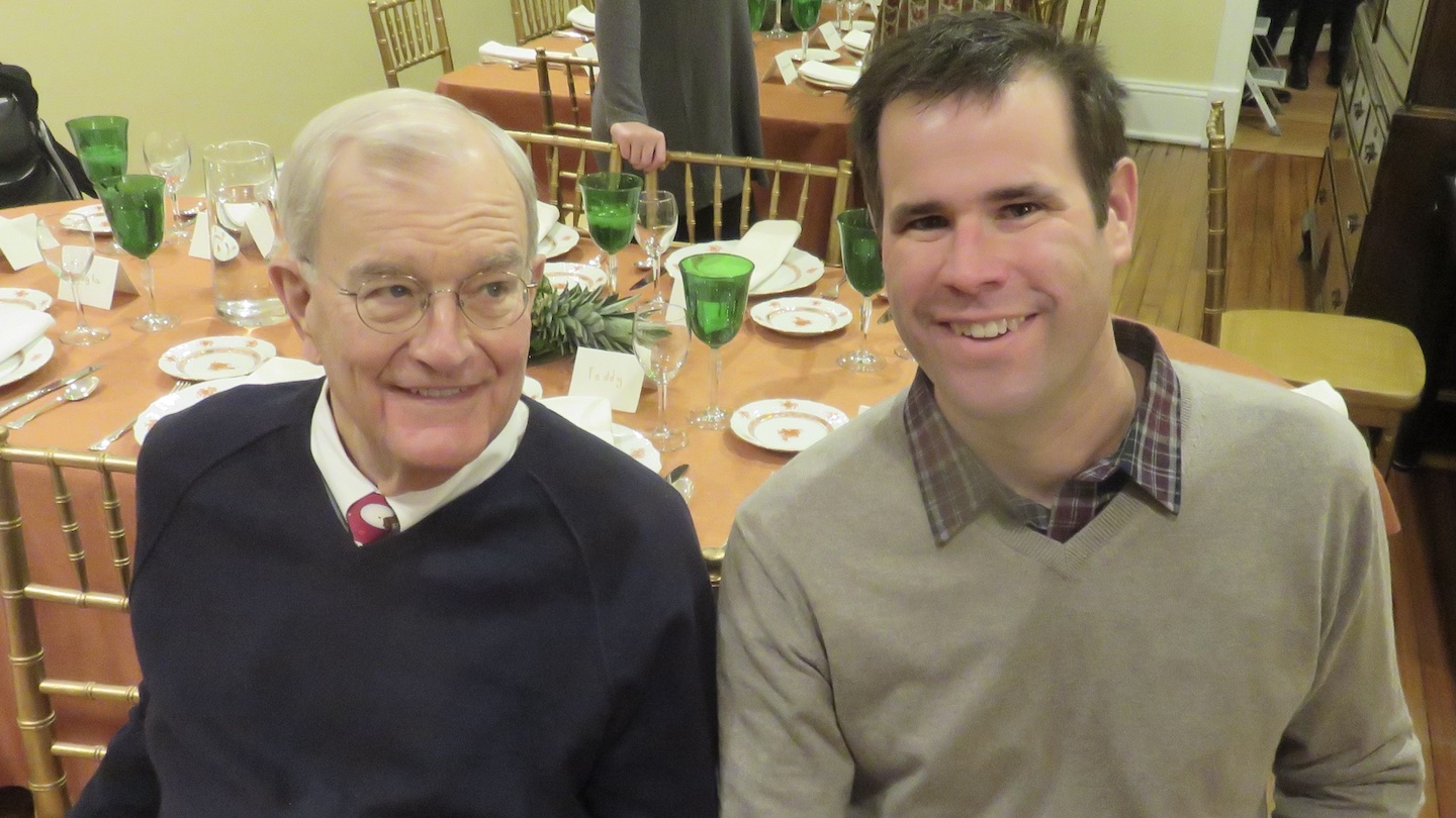 An older white man with gray hair and wireframe glasses looks to the right towards a middle aged white man with brunette hair. Both are wearing sweaters over a collared shirt. The younger man is the older man's adult son.