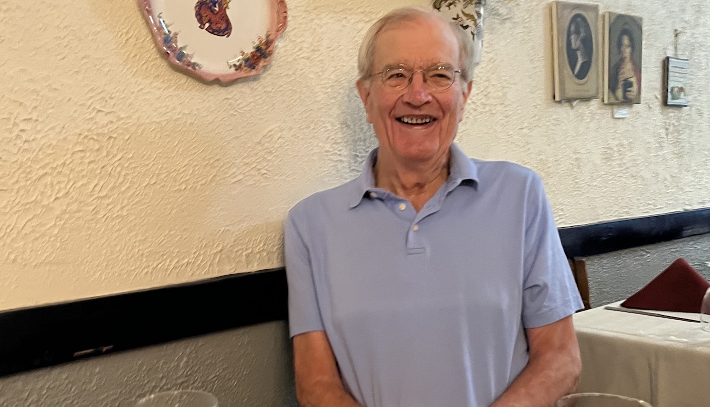 An older white man with grey hair and wire frame glasses wears a blue polo and smiles at the camera