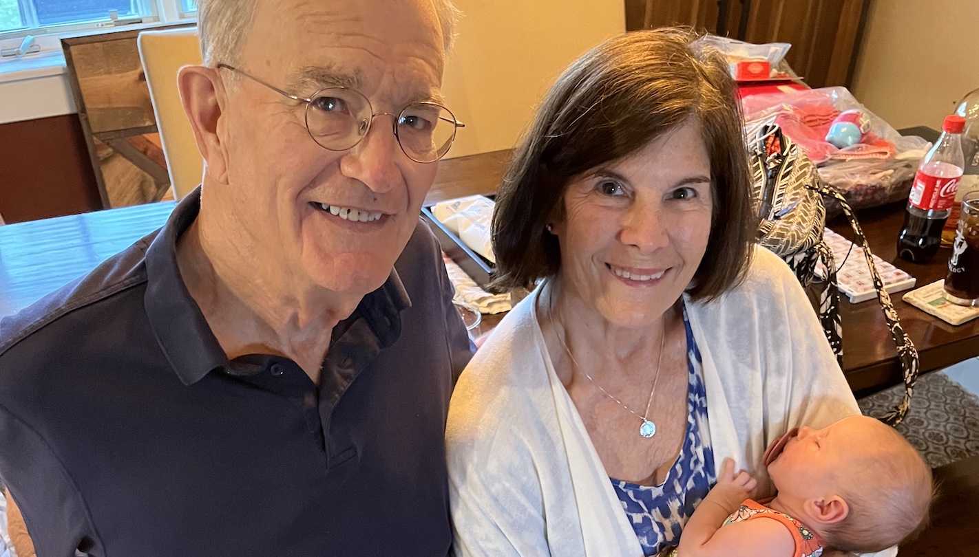 An older white man with grey hair and wireframe glasses and an older white woman with brunette hair and brown eyes both smile at the camera. The woman is holding a baby girl who is about 3 months old and sucks on a pacifier