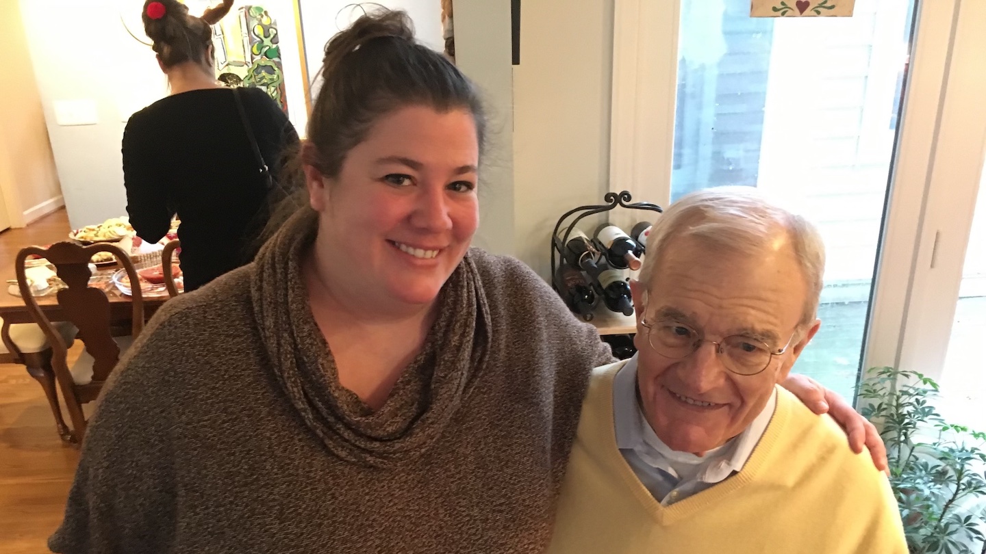 An adult woman with brunette hair and hazel eyes smiles at the camera as she sits on the lap of an older white man with gray hair and wireframe glasses. The older man is her father and he looks off camera to the right and smiles a bit awkwardly